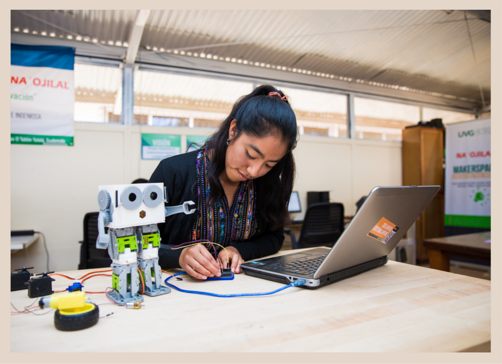 This image depicts a college-age woman dressed in clothes with patterns characteristic of one of Guatemala’s numerous indigenous communities, who is seated at a desk in a maker space at the Universidad del Valle de Guatemala. She has a laptop in front of her, and to the side of the laptop, she is conducting some electronic tests on a small robot that she has made, which is standing on the table next to her.  